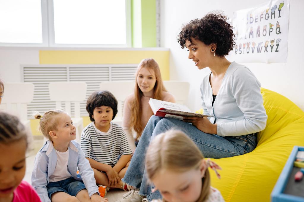 Image shows a female teacher wearing a blue button-down shirt and a blue jean trouser passionately teaching a group of five children from a book in a classroom setting.