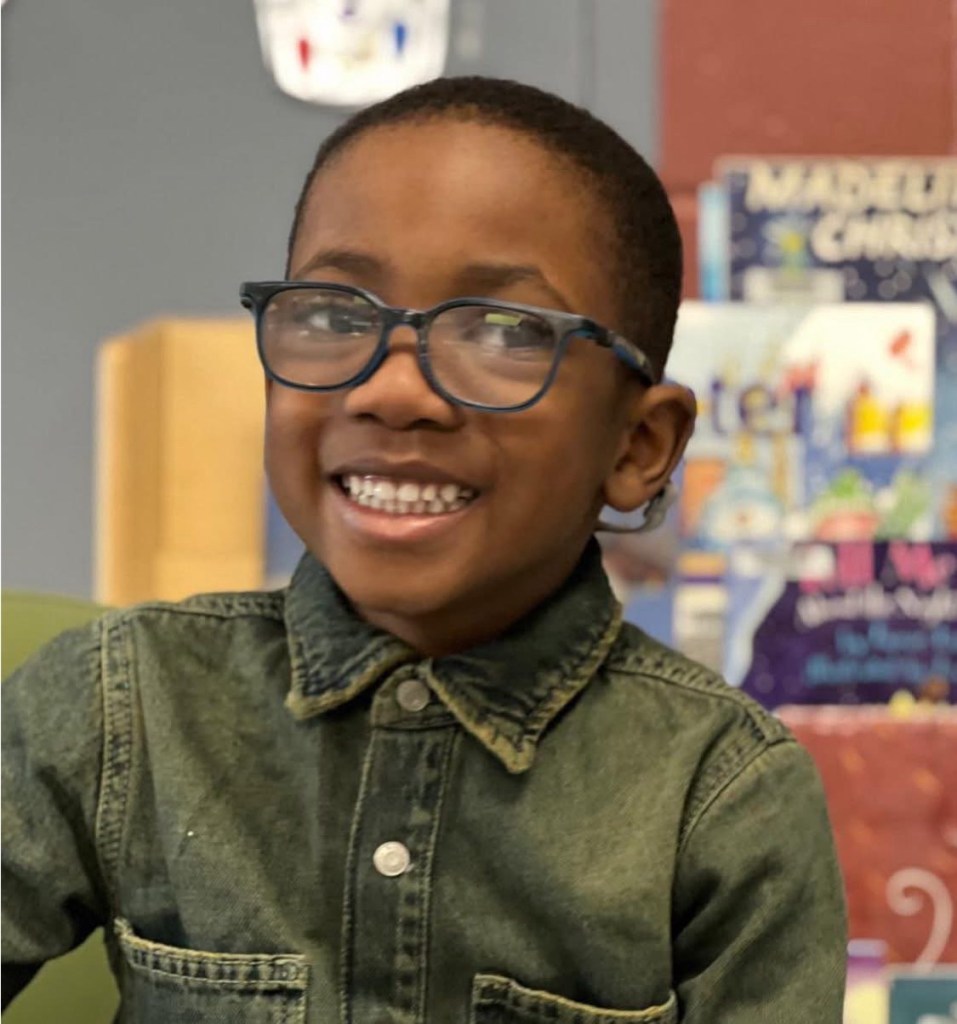 Smiling young boy wearing blue-framed eyeglasses and a buttoned-up denim shirt, seated indoors with colorful books blurred in the background.