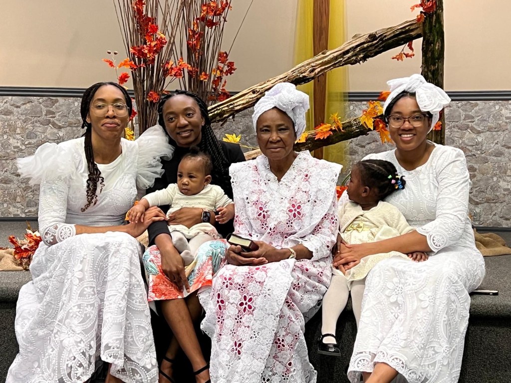 A seated portrait of three generations of women in a family — a grandmother, her daughters, and her granddaughters — dressed in white lace attire, sitting in a church decorated with autumn leaves, symbolizing faith, heritage, and spiritual legacy.