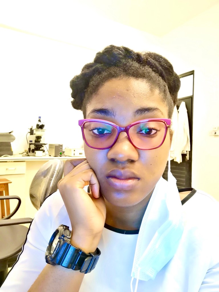  A young woman wearing red glasses and a white top sits indoors with her chin resting on her hand. She has natural hair styled in a twisted updo, a wristwatch on her left hand, and a face mask hanging loosely from one ear. She looks thoughtful, with lab equipment visible in the background.