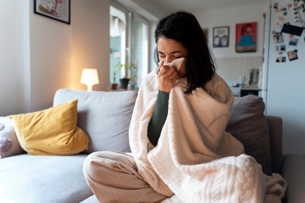 Woman sitting on a sofa under a blanket, holding a tissue to her face while unwell at home, symbolizing moments of weakness and the comfort of care.