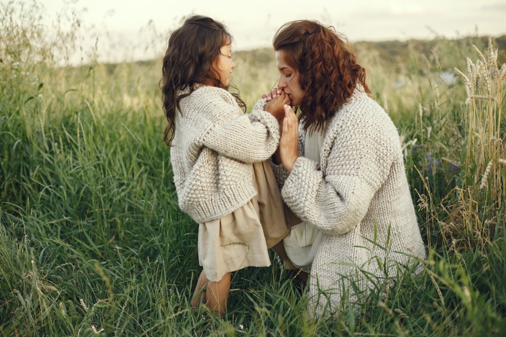 A mother kneels in a grassy field, gently holding her daughter’s hand while they wear matching knitted sweaters — symbolizing legacy, nurturing, and passing wisdom to the next generation.